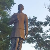 A dark bronze statue of Gurram Jashuva in glasses and a long buttoned coat, holding a rolled paper, on a red granite plinth with a black Telugu plaque, among trees