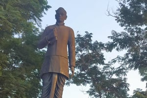 A dark bronze statue of Gurram Jashuva in glasses and a long buttoned coat, holding a rolled paper, on a red granite plinth with a black Telugu plaque, among trees