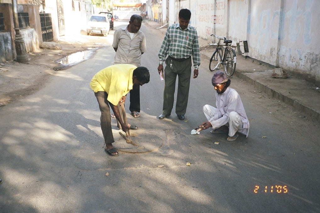 On a street in Ahmedabad, one man levers up a round sewer cover with an iron rod while another crouches beside it and two more stand by