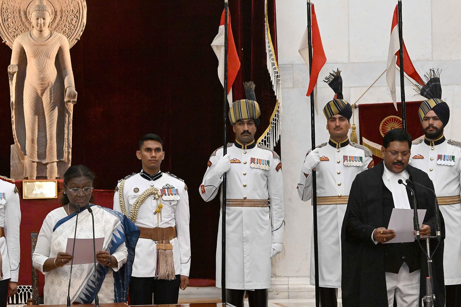 B.R. Gavai, in black judicial robes, reads the oath at a microphone while President Droupadi Murmu reads beside him, ceremonial guards standing behind