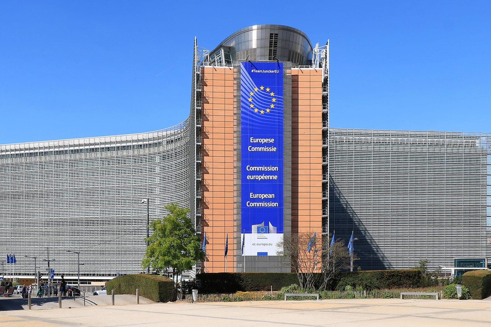 The Berlaymont's curved glass facade beneath a blue European Commission banner and a clear sky