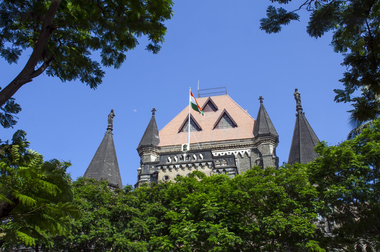 The pointed turrets and flag of the Bombay High Court in Mumbai above green trees against a blue sky