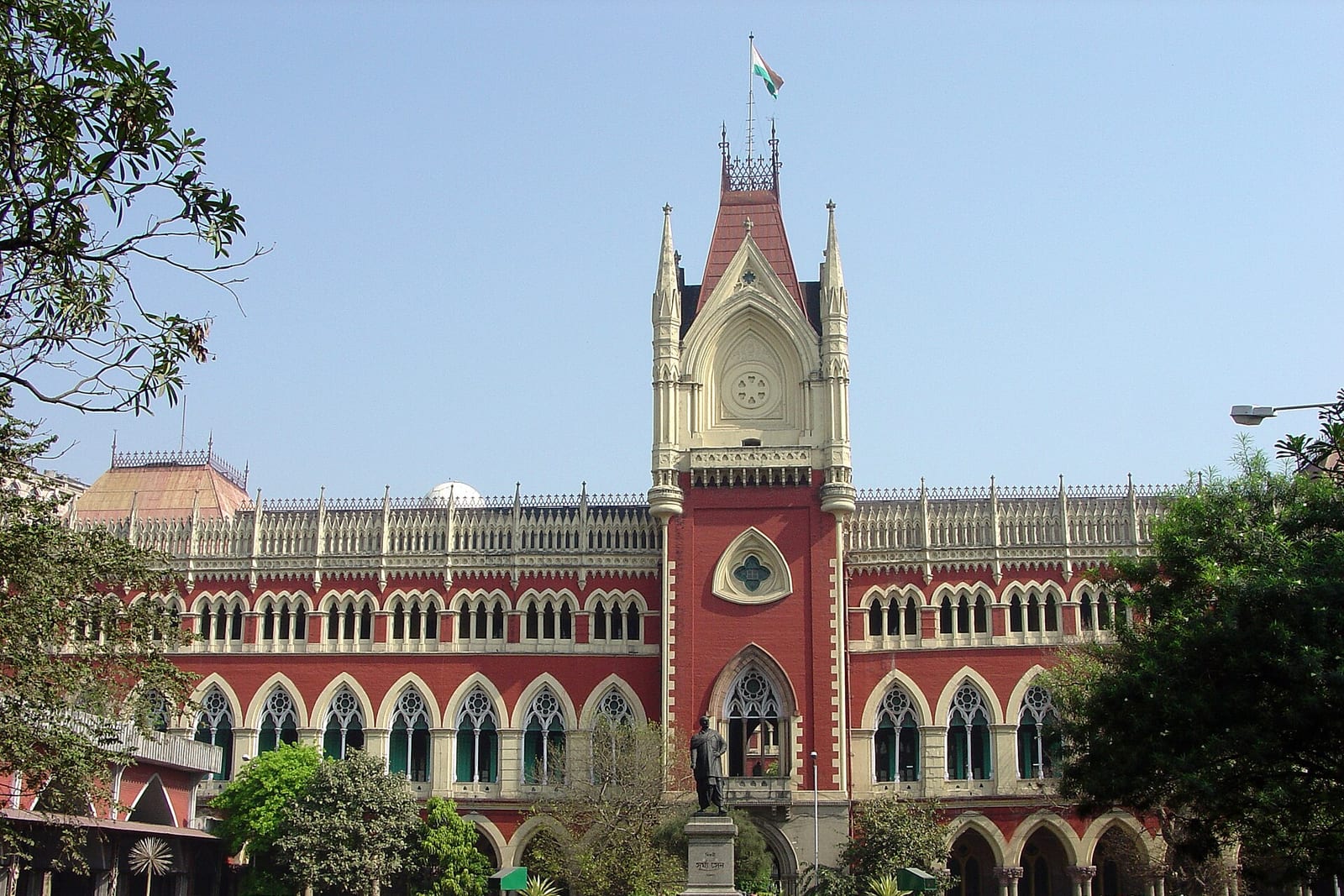 The Calcutta High Court building in Kolkata, a red-and-cream Gothic facade with a central clock tower, seen from the street in daylight
