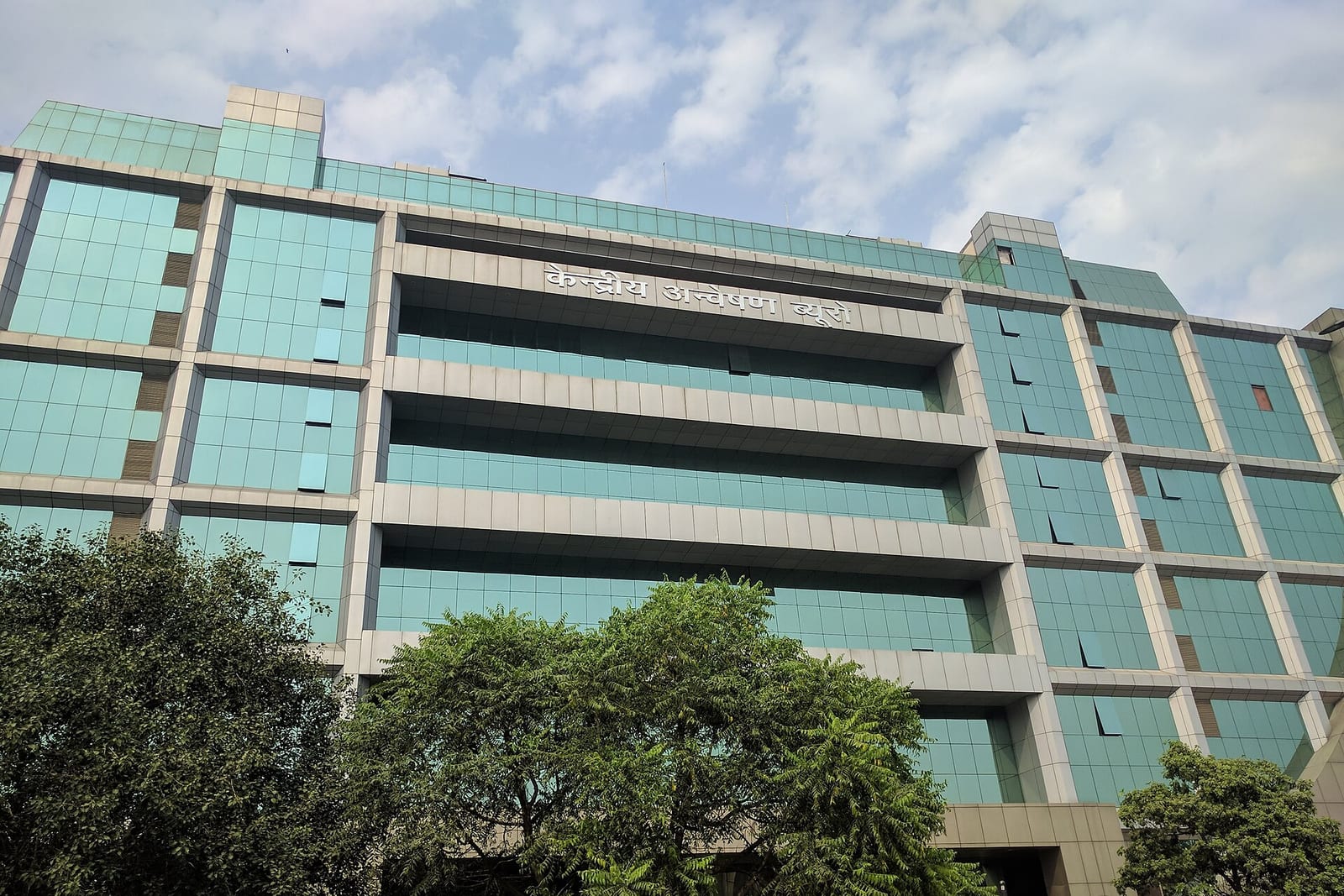 A multi-storey office building with green-tinted glass and Hindi lettering naming the Central Bureau of Investigation, behind trees