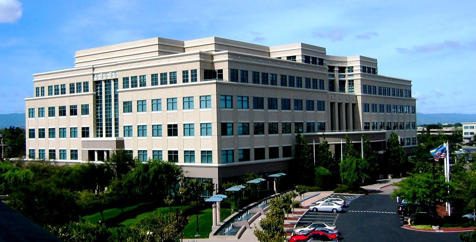 A pale six-storey office building with rows of green-tinted windows, trees and a car park in front, under a blue sky