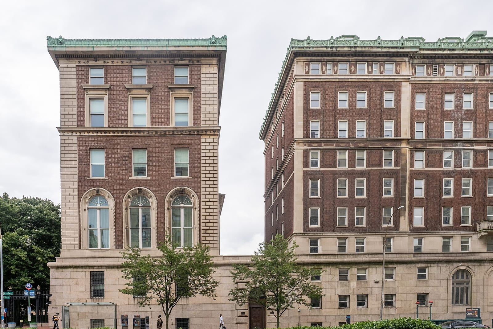 The brick and limestone facade of Pulitzer Hall and a taller neighbouring Columbia building on Broadway, with young street trees and a few passers-by on the pavement