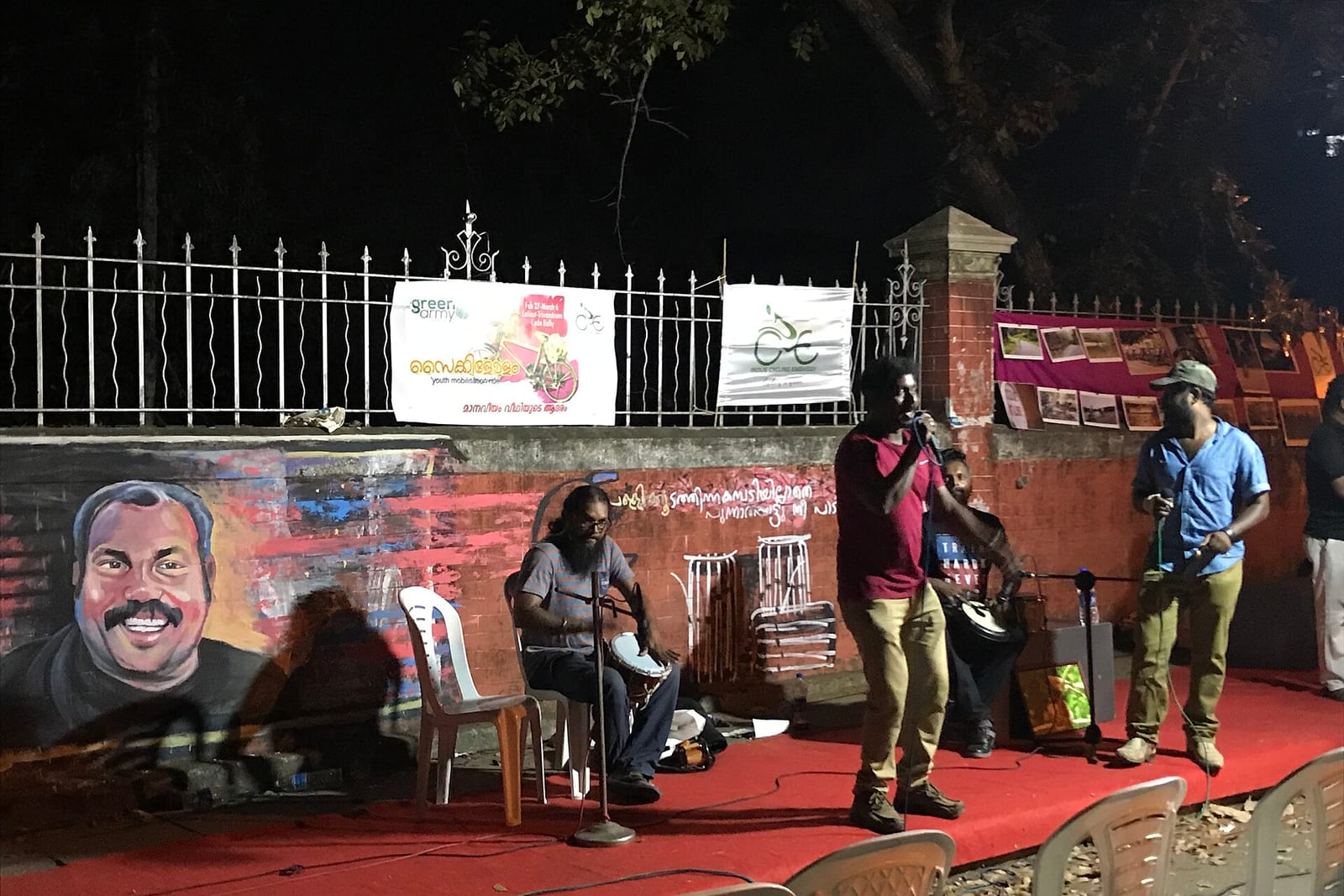 Musicians perform at night on a red-carpeted pavement stage beside a painted wall mural of a smiling Kalabhavan Mani