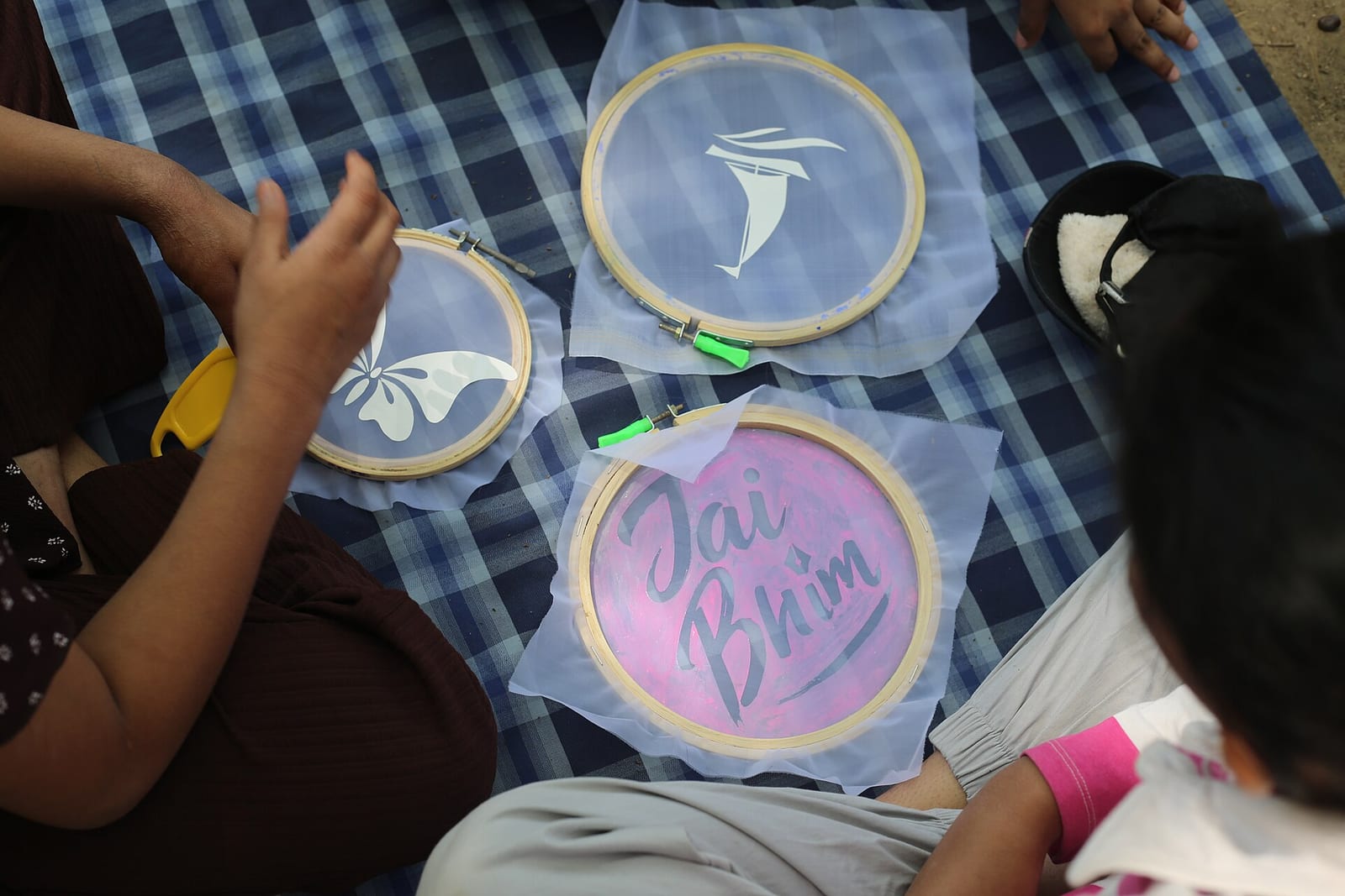 Hands working on embroidery-hoop screens on a blue checked mat; one screen is printed with the words Jai Bhim, an Ambedkarite greeting, others with a white butterfly and a sailing-boat shape