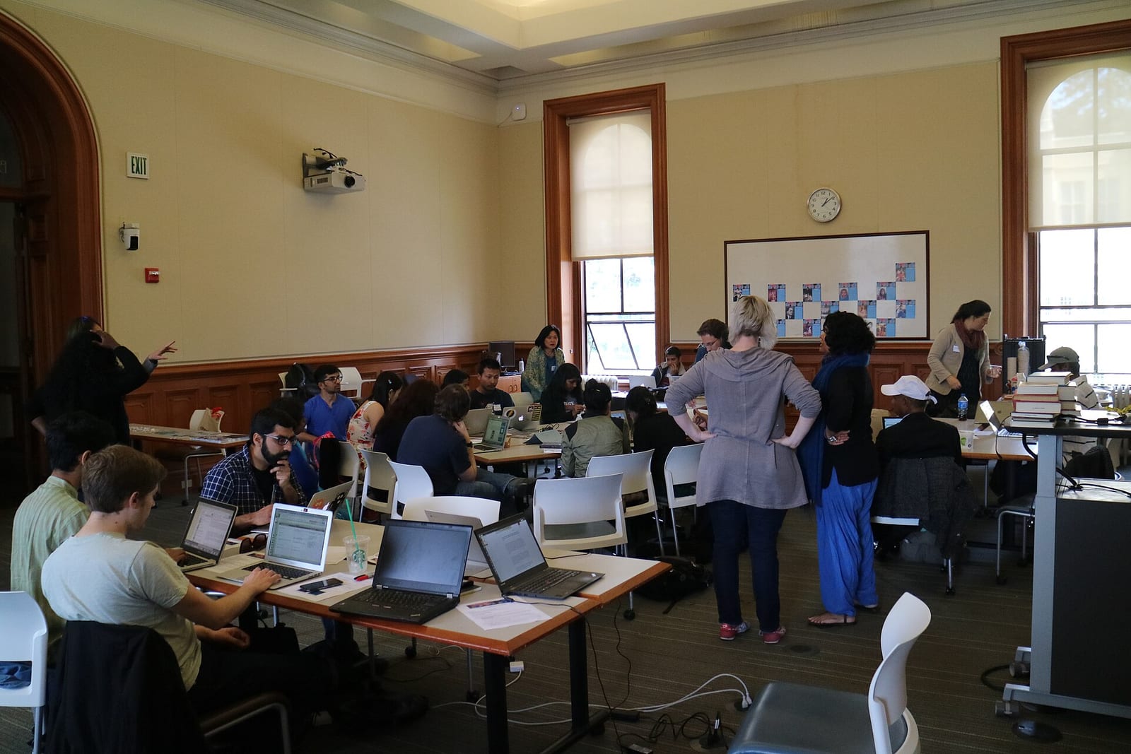 About two dozen people work on laptops at long tables in a wood-panelled room, some standing and talking