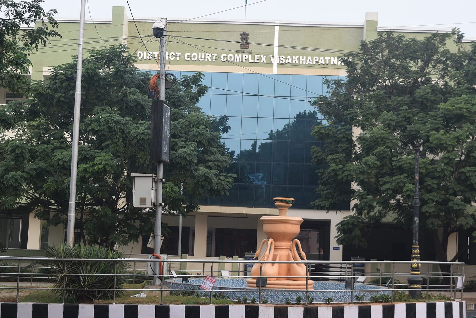 The glass-fronted District Court Complex in Visakhapatnam, behind trees and a roadside railing