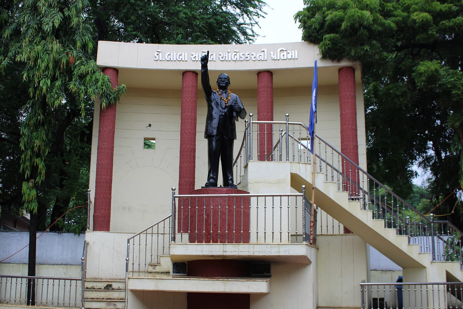 A dark bronze statue of B.R. Ambedkar with a raised right arm and a marigold garland, on a pedestal in front of a cream pavilion with red pillars, beside a blue flag