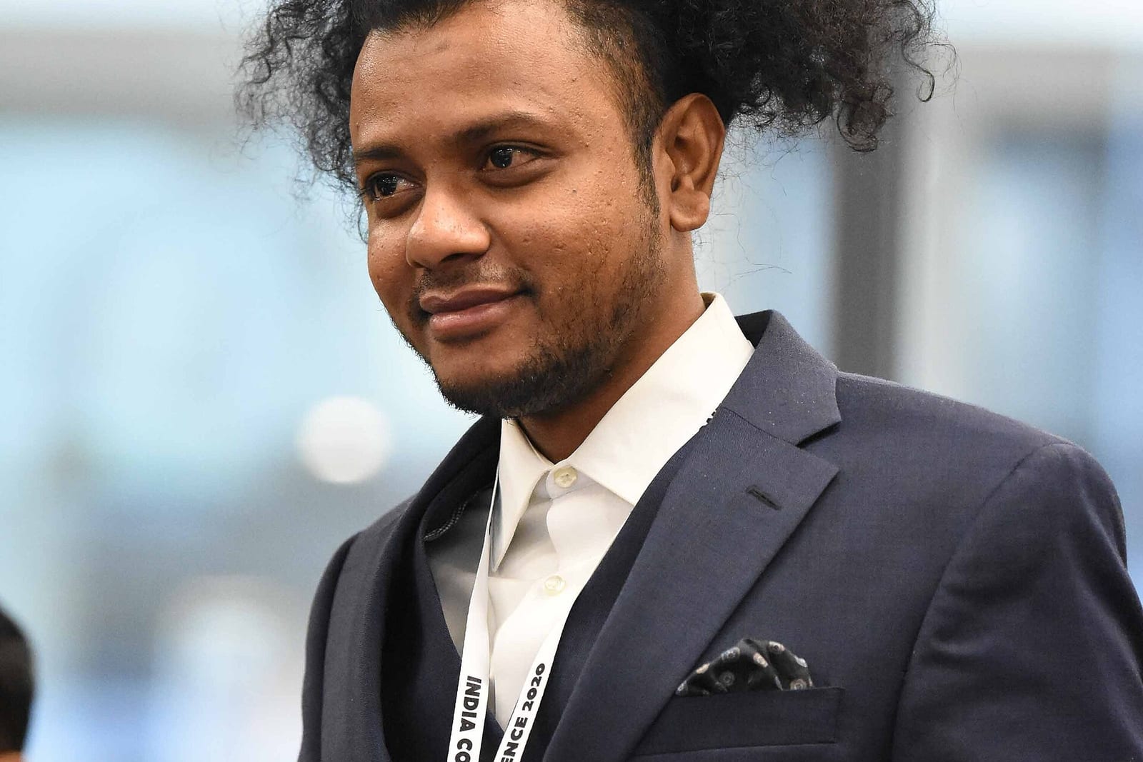 Suraj Yengde, a man with voluminous curly hair and a short beard, in a navy suit and white shirt with a conference lanyard, smiling slightly
