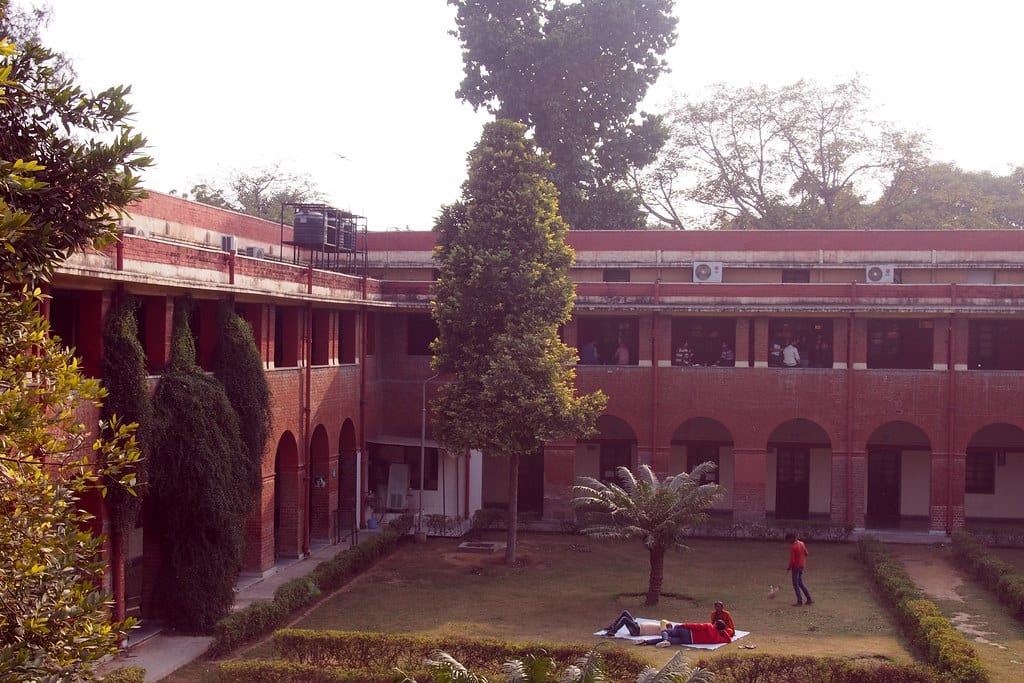 A red-brick building with arched colonnades around a grassy courtyard; a few people are visible in the distance
