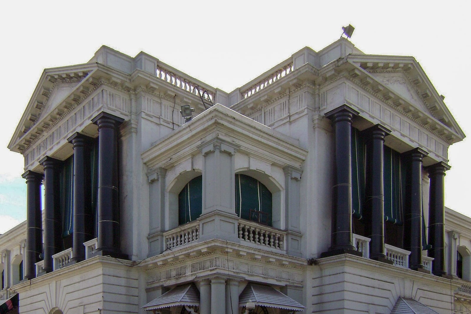 A white colonial-era building with tall black columns, balustrades and green window blinds, seen from a corner under a pale sky