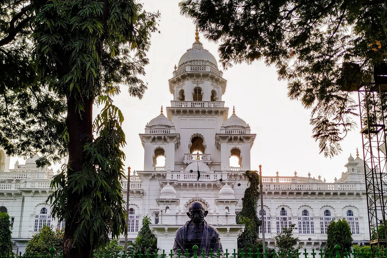 The white, domed Telangana Legislative Assembly building in Hyderabad, seen through trees, with a statue of Mahatma Gandhi in front