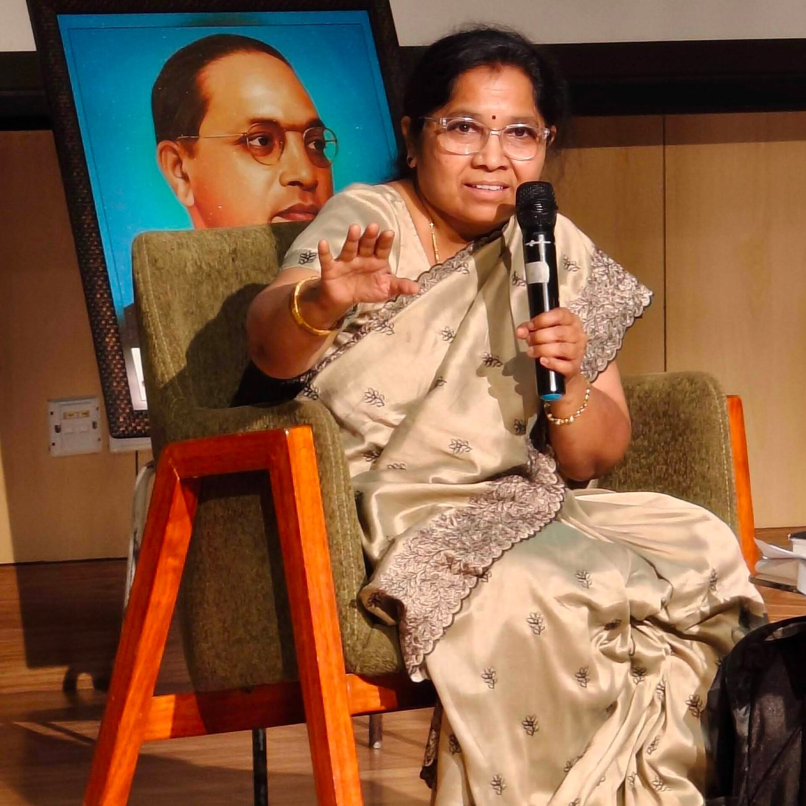 Gogu Shyamala, in a cream silk sari and glasses, speaks into a microphone from an armchair, one hand raised, with a framed portrait of B.R. Ambedkar behind her