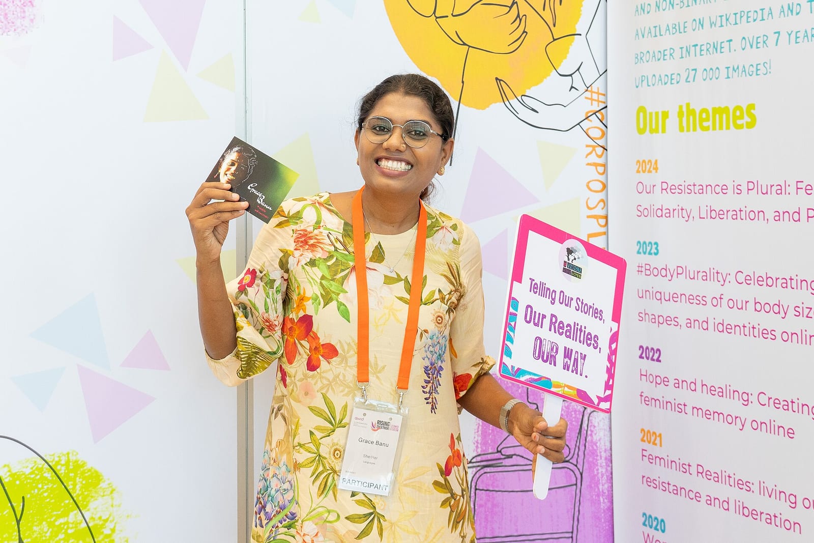 Grace Banu smiles, holding up a postcard and a sign reading 'Telling Our Stories, Our Realities, Our Way'