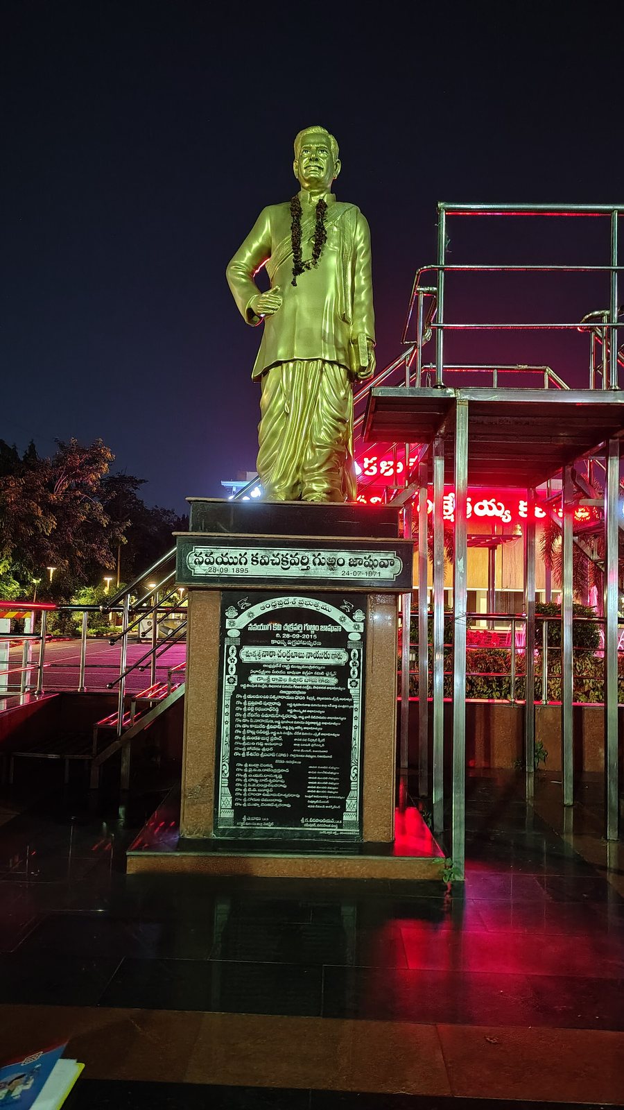 A gilded, garlanded statue of Gurram Jashuva on a stone plinth, lit at night