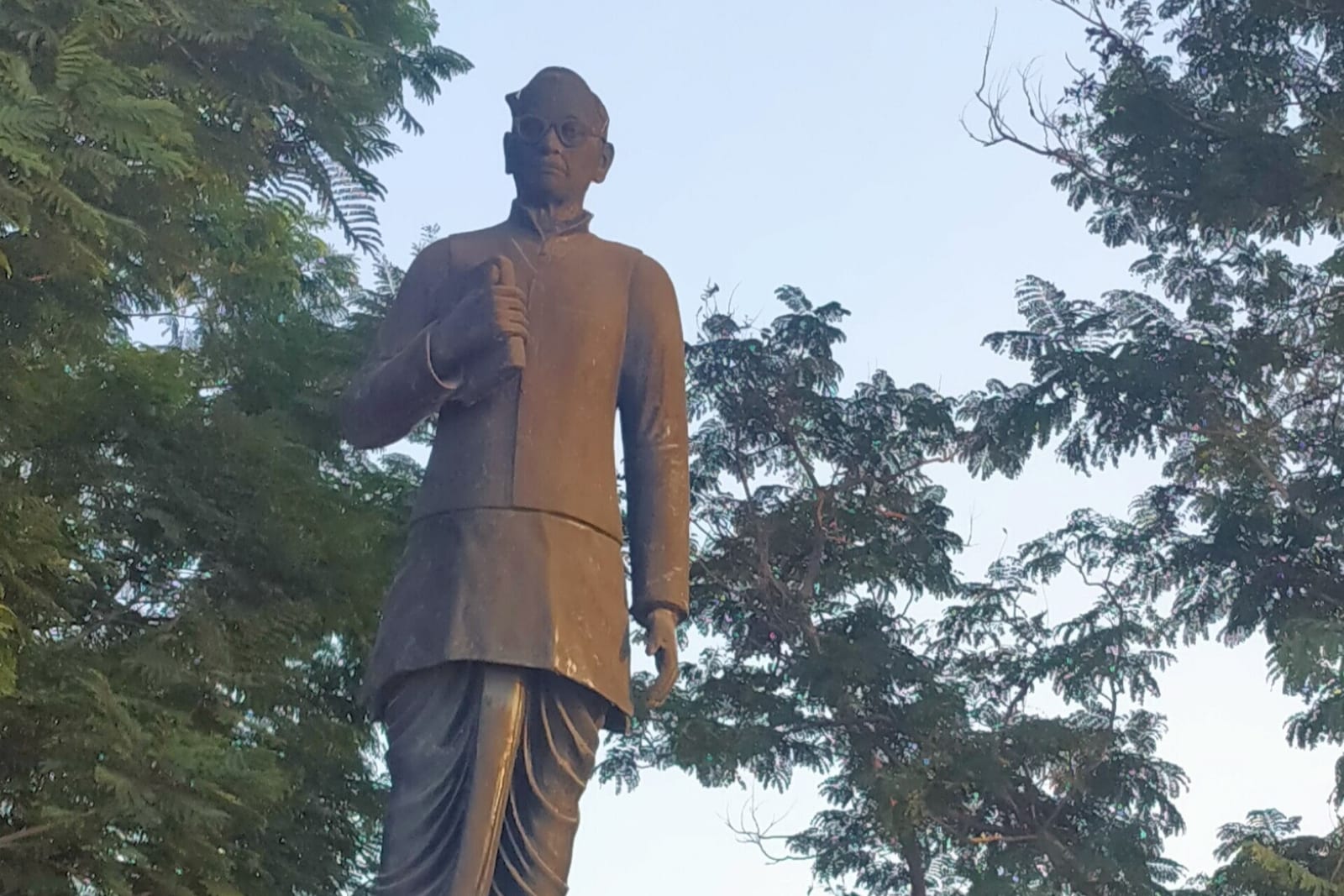 A dark bronze statue of Gurram Jashuva in glasses and a long buttoned coat, holding a rolled paper, on a red granite plinth with a black Telugu plaque, among trees