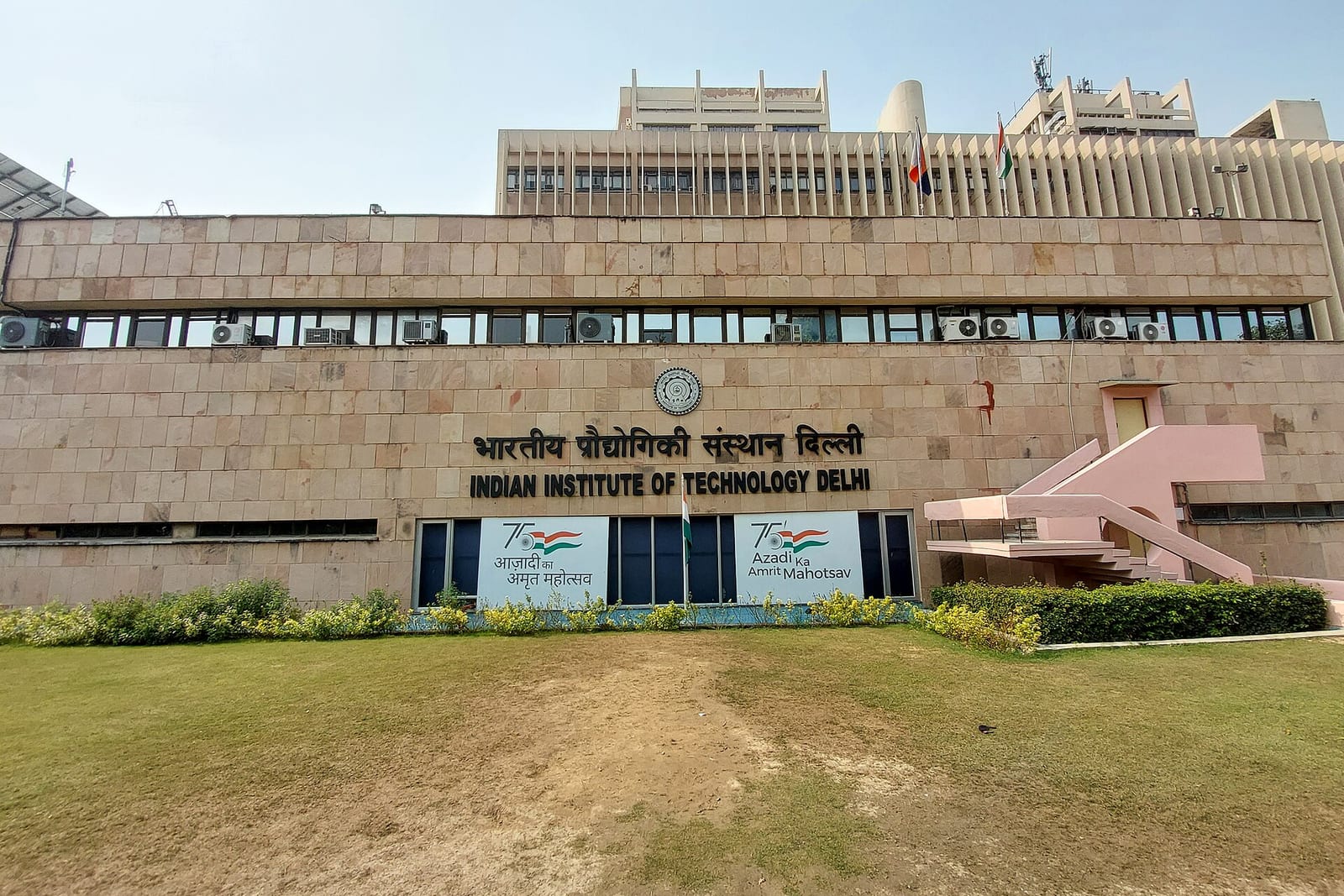 The stone-clad main building of IIT Delhi, its name in Hindi and English above the entrance, behind a lawn