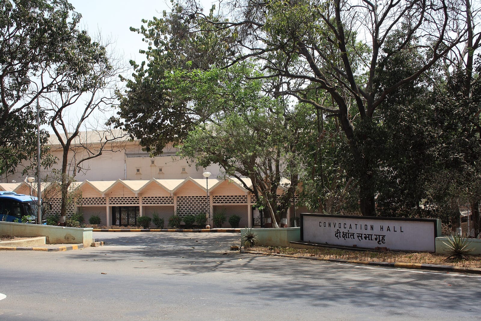 A low pale building with a scalloped roof behind trees, with a sign reading Convocation Hall in English and Hindi