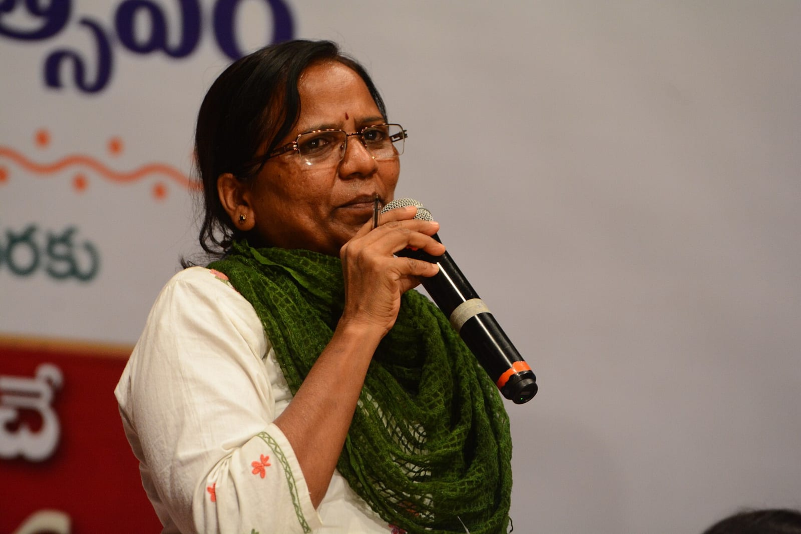 Joopaka Subhadra, in glasses, a white kurta and a green scarf, speaks into a microphone on a stage