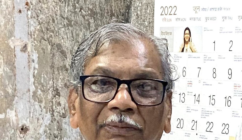 J.V. Pawar, an older man with grey hair and black-rimmed glasses, in front of a wall calendar