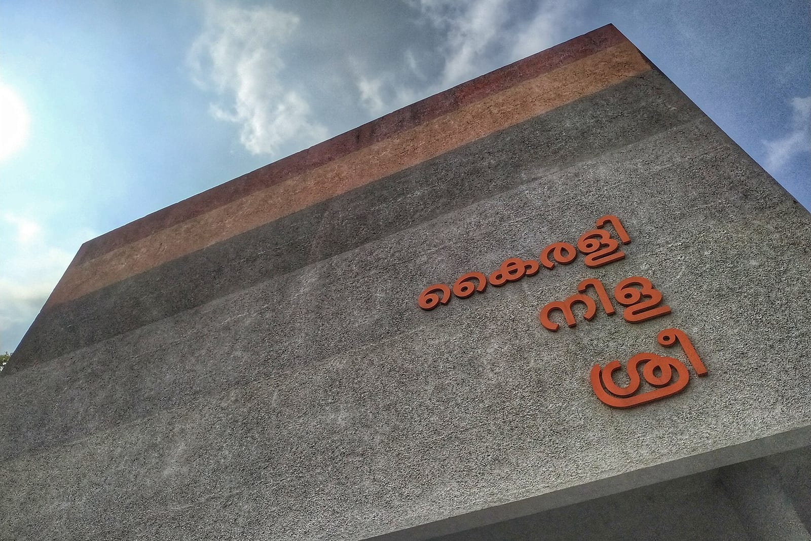 Orange Malayalam lettering naming the Kairali, Nila and Sree theatres on a grey concrete wall, seen from below against a blue sky