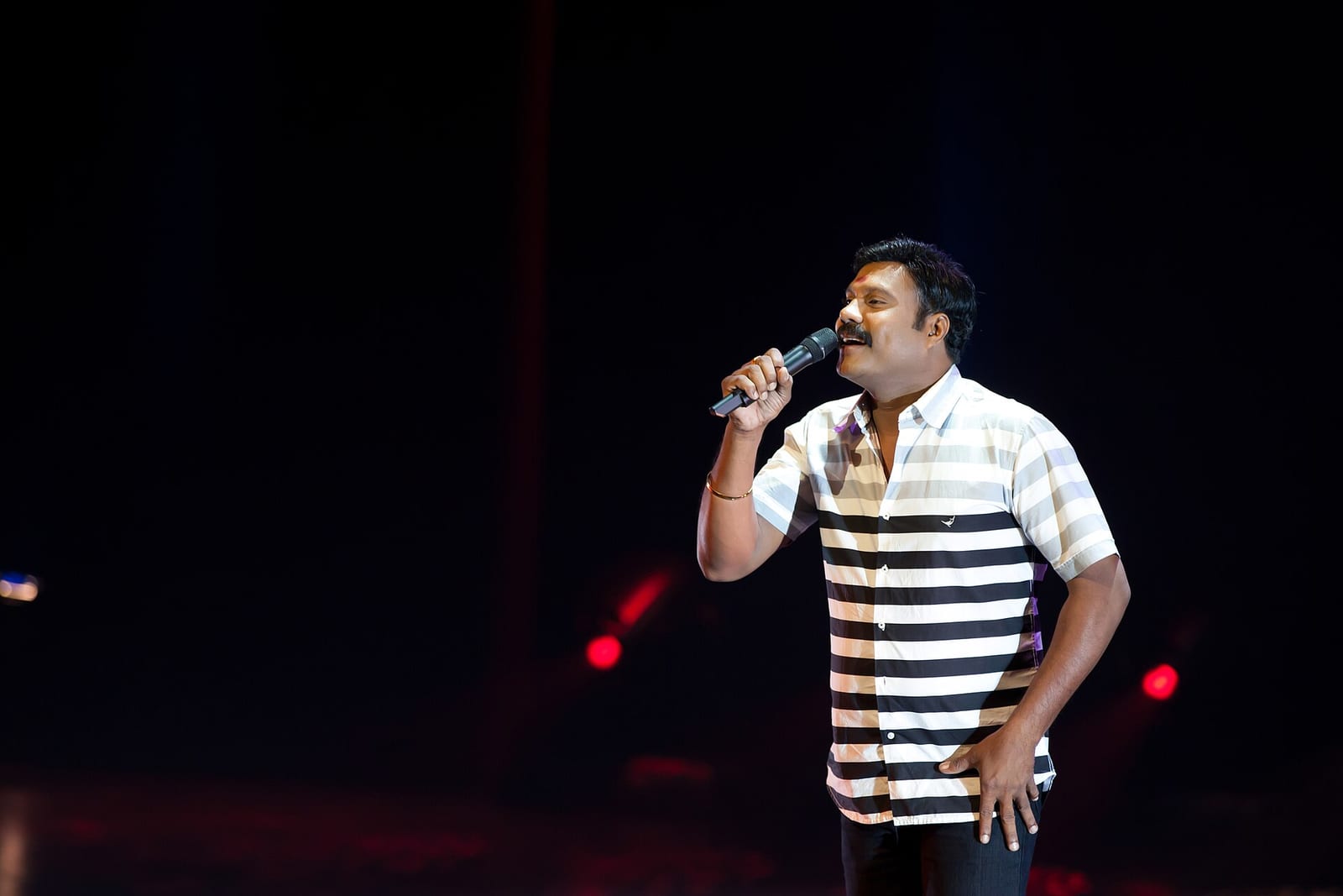 Kalabhavan Mani, in a black-and-white striped shirt, sings into a microphone on a dark stage