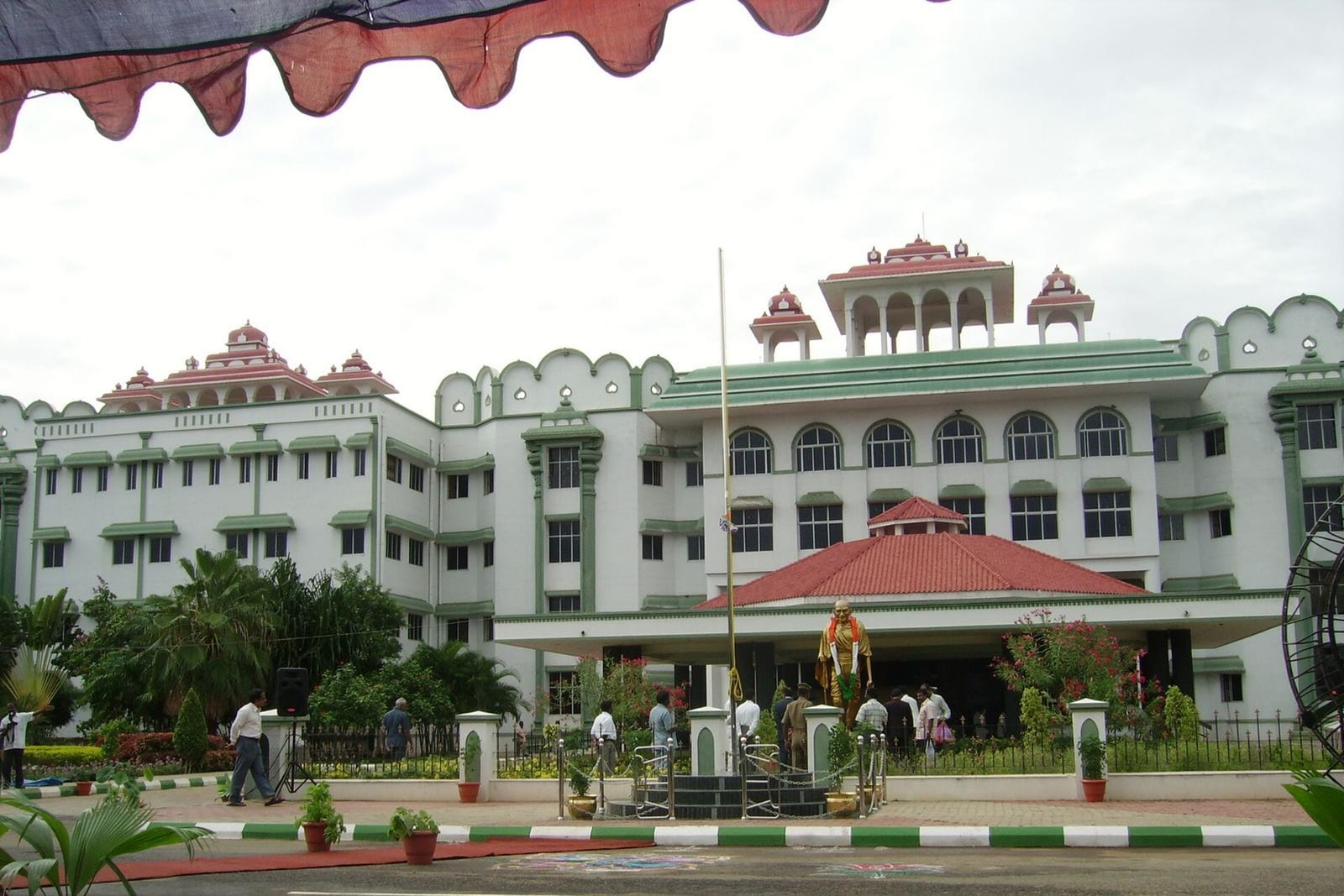 A large white building with green trim and red-tiled rooftop pavilions, with a flagpole and a statue in front of its entrance
