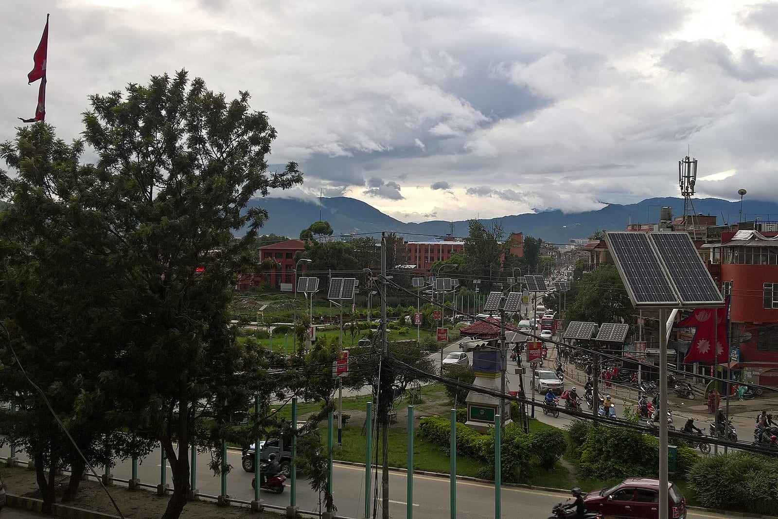 Traffic circles a green roundabout with a small monument in central Kathmandu, under cloudy skies with hills behind