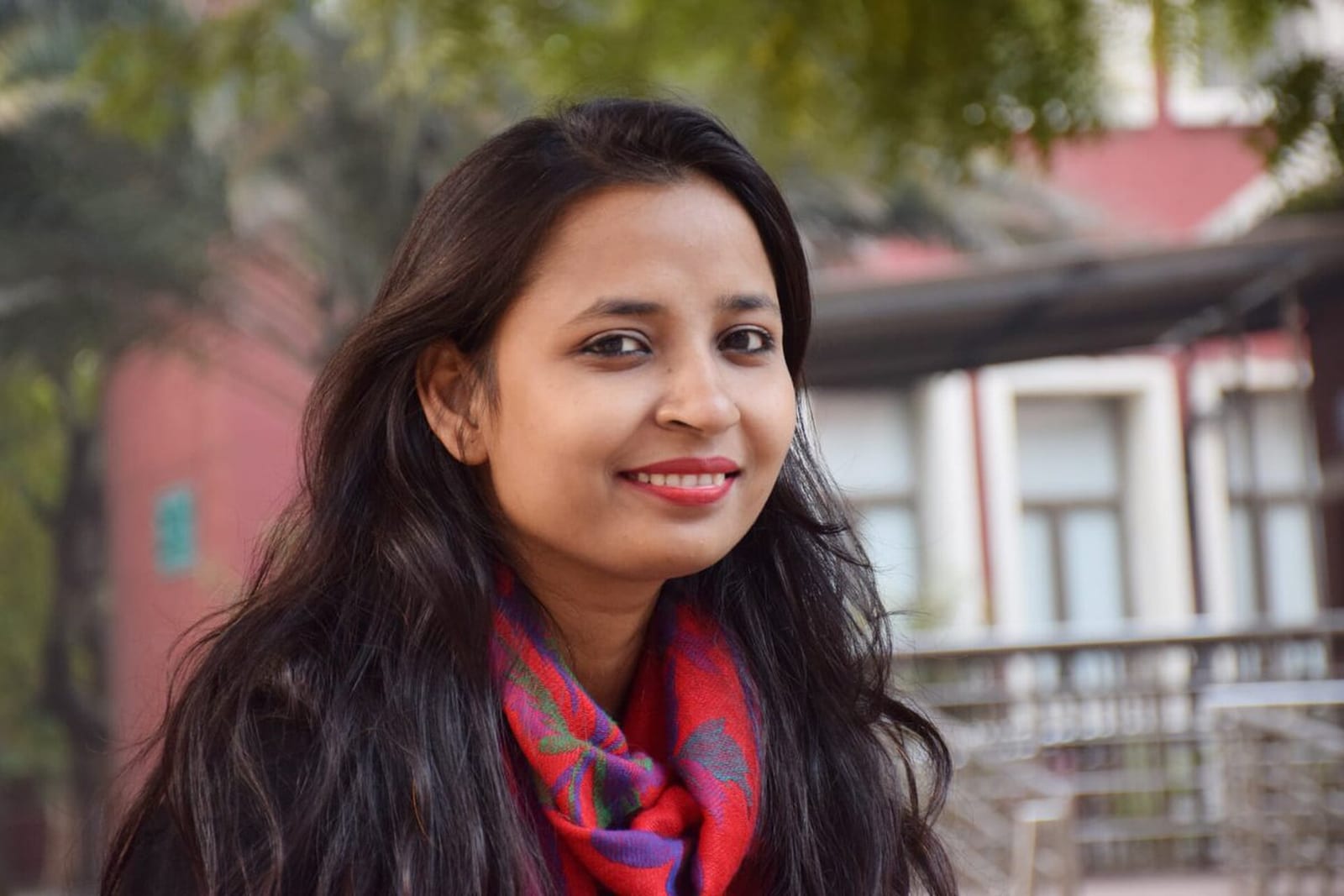 Meena Kotwal, smiling, with long dark hair and a red and purple scarf, outdoors in front of a red-brick building