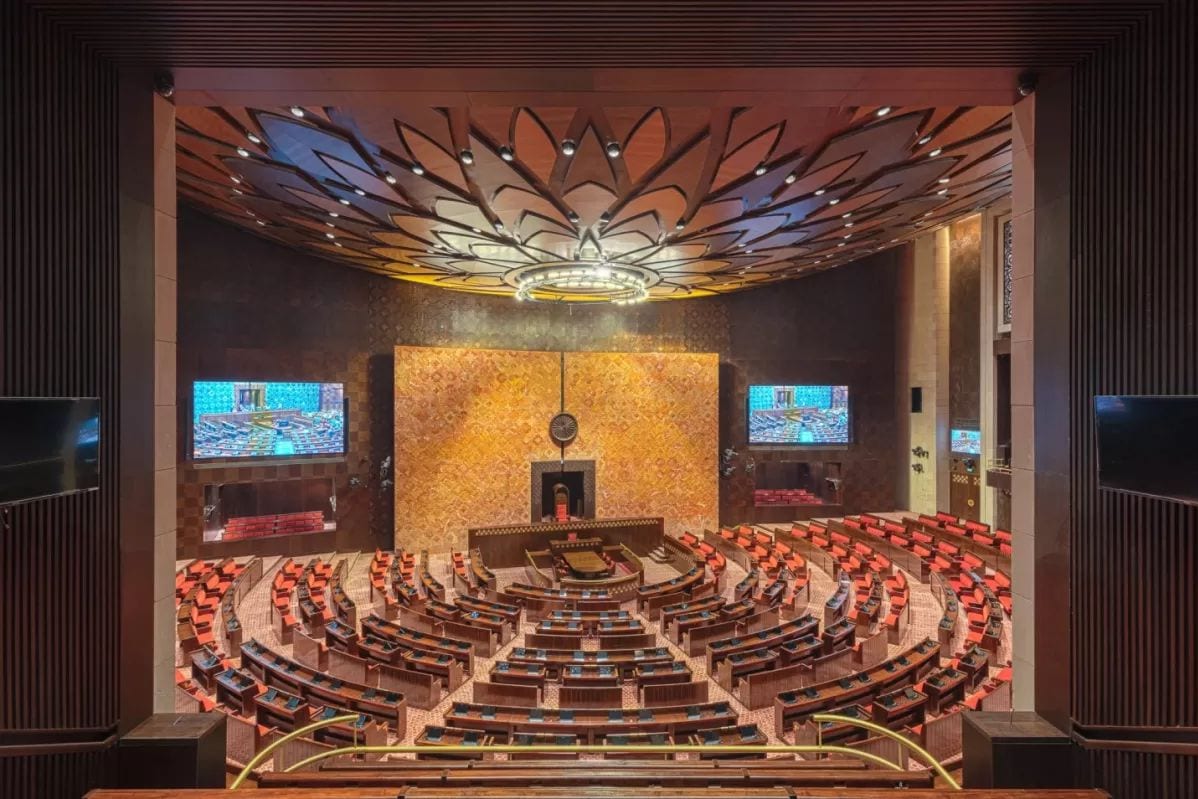 An empty legislative chamber with rows of red seats arranged in a horseshoe around the presiding officer's chair, under a lotus-patterned ceiling