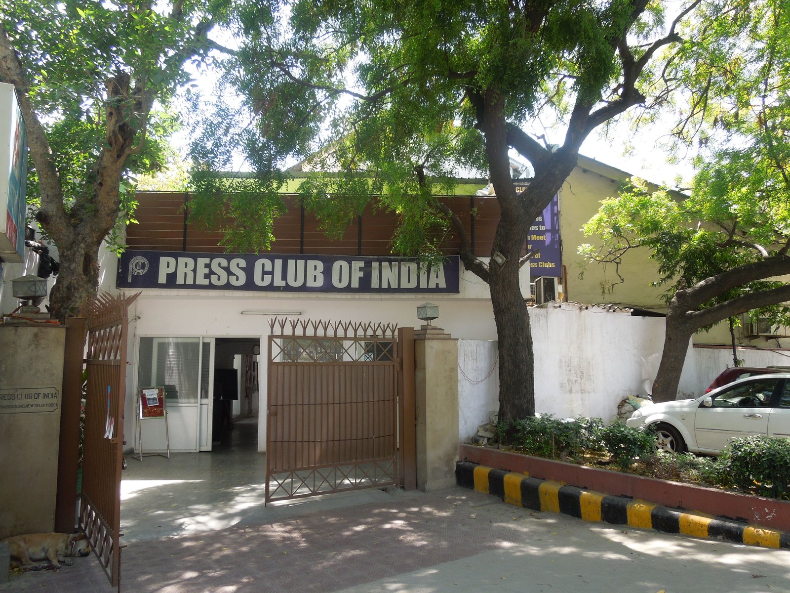 The gated entrance of the Press Club of India, a low white building under trees with a dark blue name board