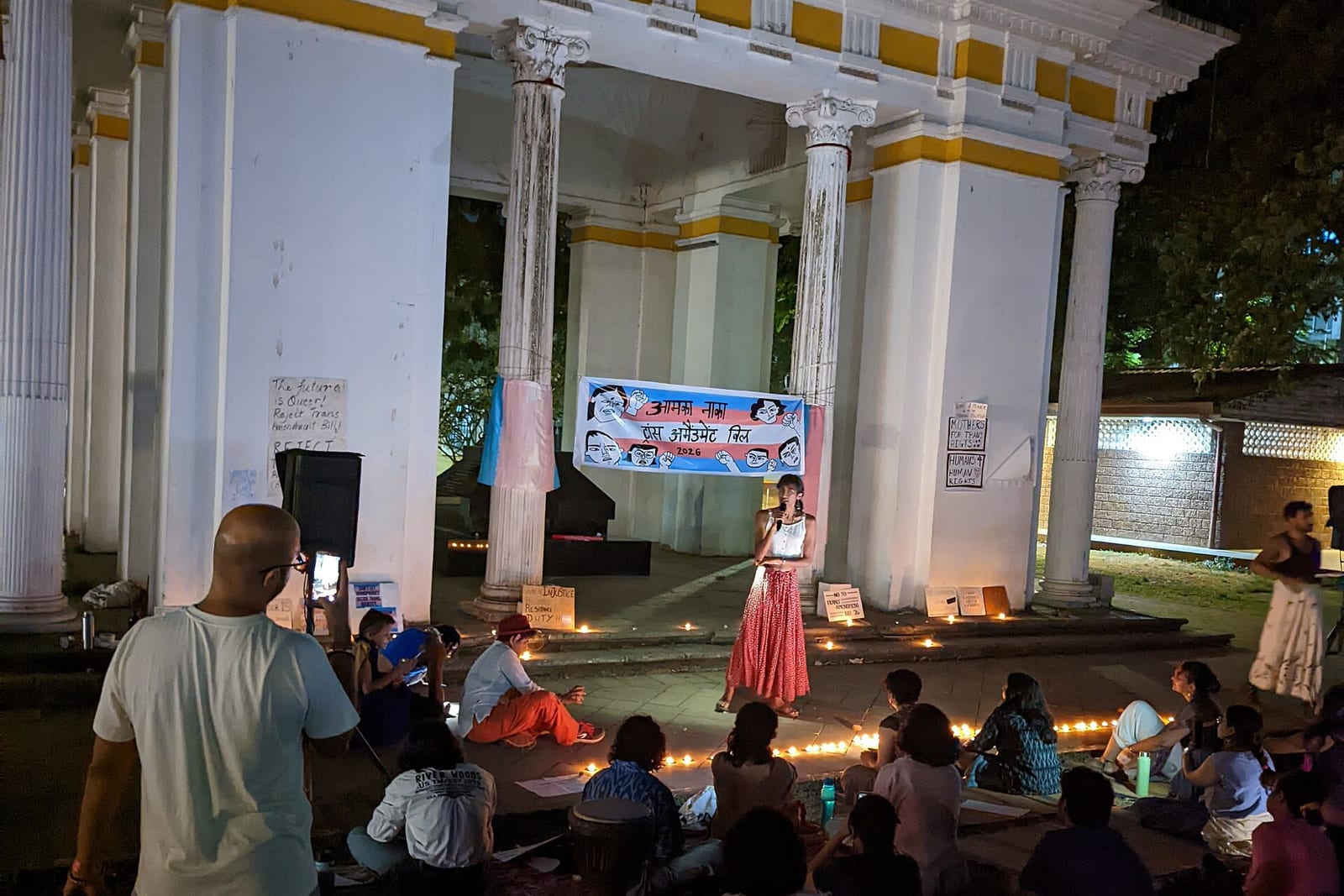 People sit among candles at night before a white colonnaded pavilion as a speaker addresses them beneath a protest banner