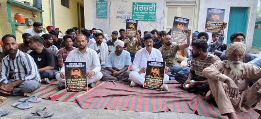 Men sit on mats outside the Sangrur civil hospital mortuary holding posters reading Justice for Sanamdeep