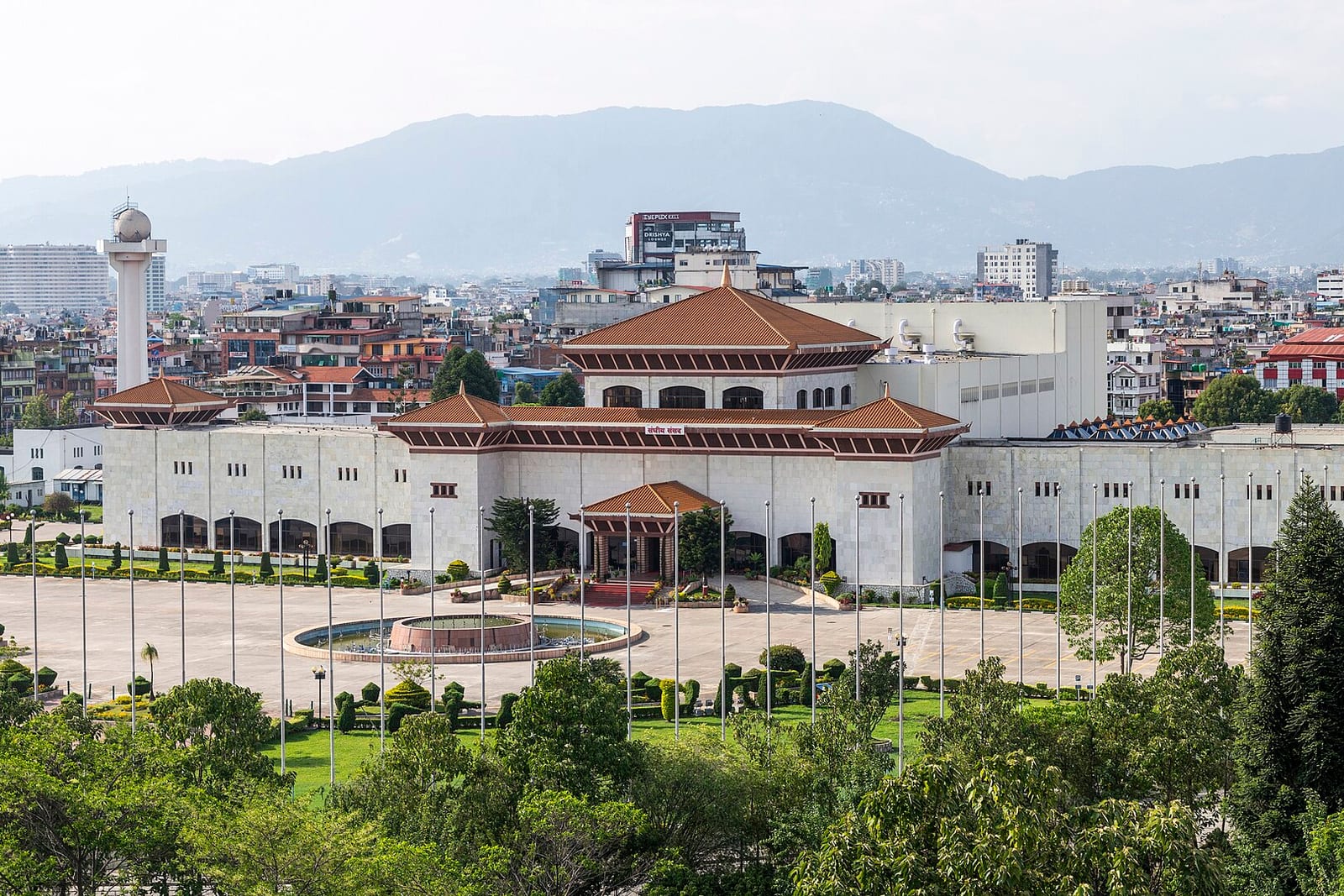 Nepal's Federal Parliament building, a long white complex with tiered terracotta roofs and a flagpole-lined forecourt, with Kathmandu's hills behind