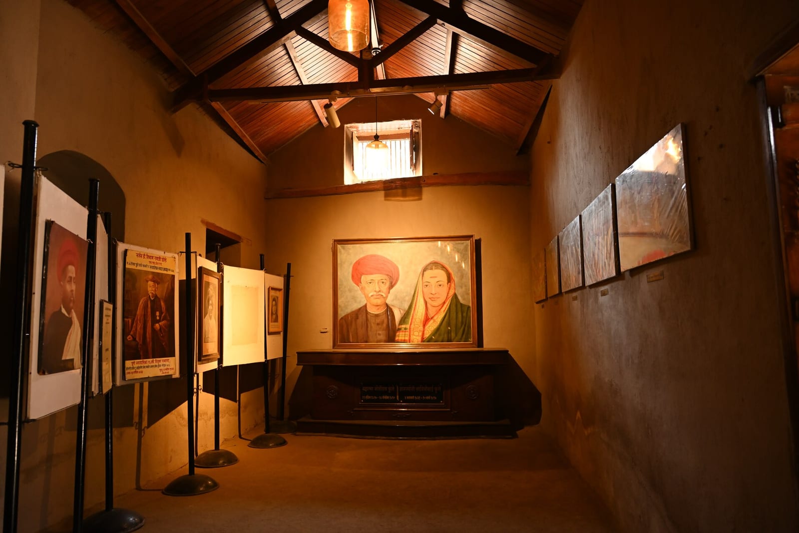 A painted portrait of Jyotirao and Savitribai Phule hanging in a dim room with display boards, at Phulewada in Pune