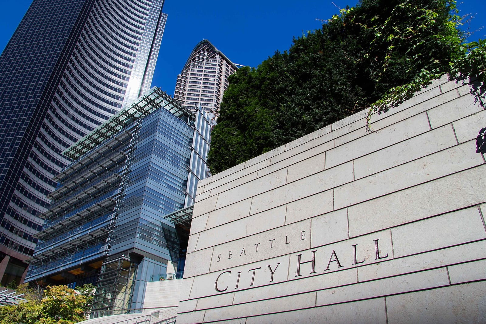 The words Seattle City Hall carved into a pale stone wall, with glass towers and a blue sky behind