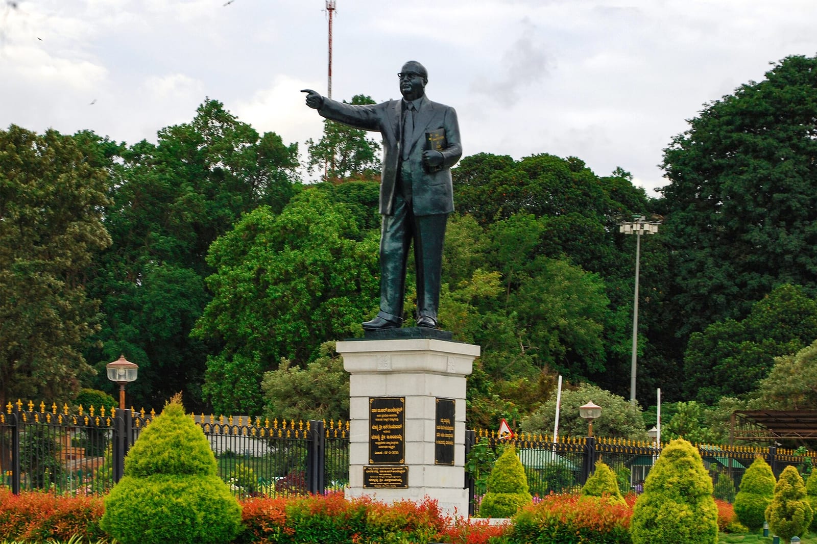 A dark statue of B.R. Ambedkar in a suit, one arm pointing and a book in the other hand, on a white plinth among trimmed hedges and trees