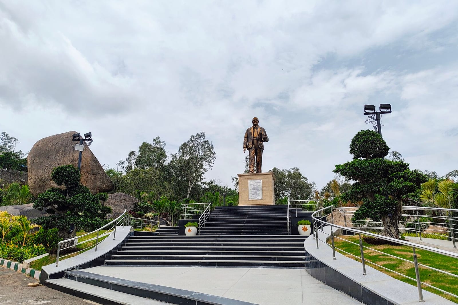 A bronze statue of B.R. Ambedkar on a pedestal at the top of wide stone steps, among trees and boulders, under a cloudy sky