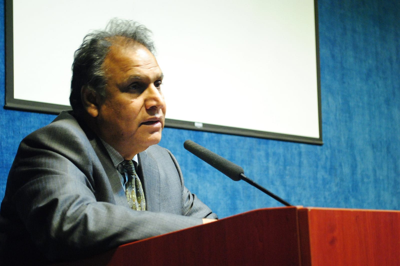 Sukhadeo Thorat, in a grey suit, speaks at a lectern in front of a blank projector screen