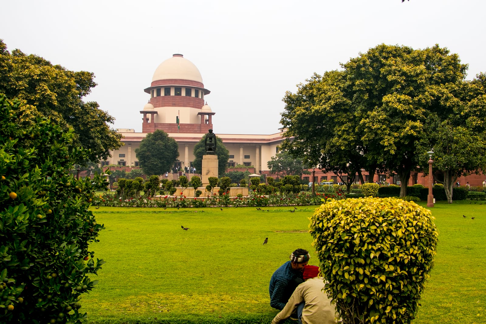 The domed central wing of the Supreme Court of India behind a lawn and trees, with two gardeners at work in the foreground