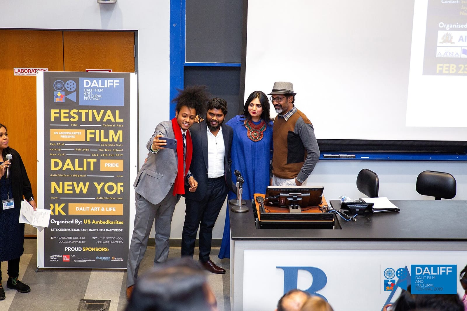 Four people pose for a selfie in front of a projector screen beside a banner for the Dalit Film and Cultural Festival, New York