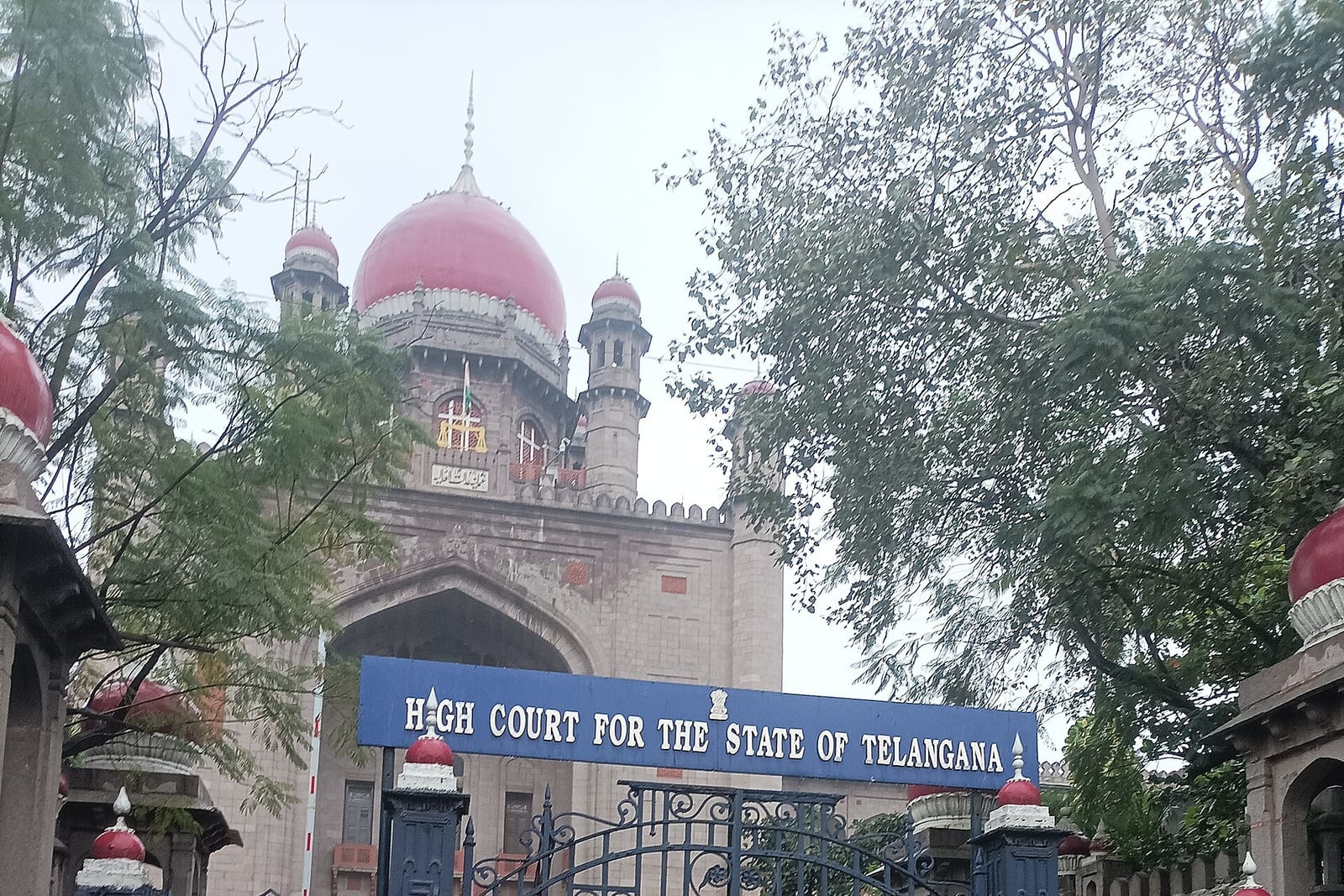 The domed entrance of the High Court for the State of Telangana in Hyderabad, seen through trees, with its blue name board over the gate.