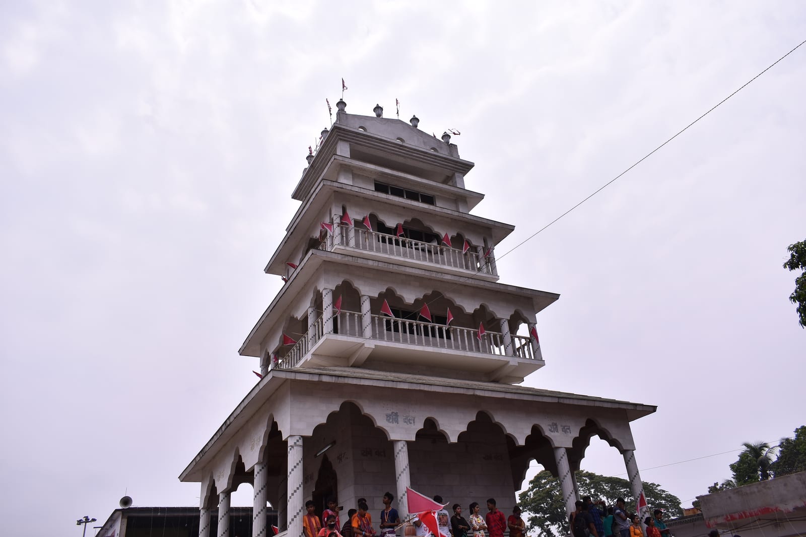 The white, multi-tiered tower of the Thakurbari temple at Thakurnagar, hung with small red flags, with visitors gathered at its base