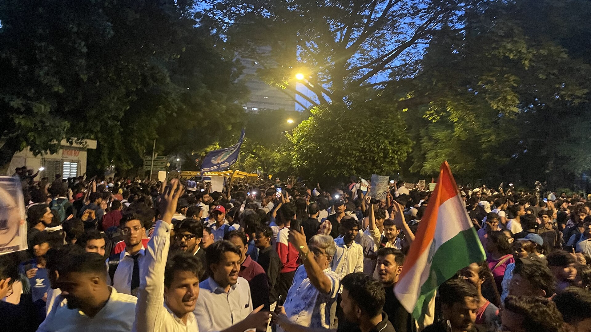 A crowd of young protesters at night at Jantar Mantar, Delhi, under street lights, with a blue flag and an Indian flag raised