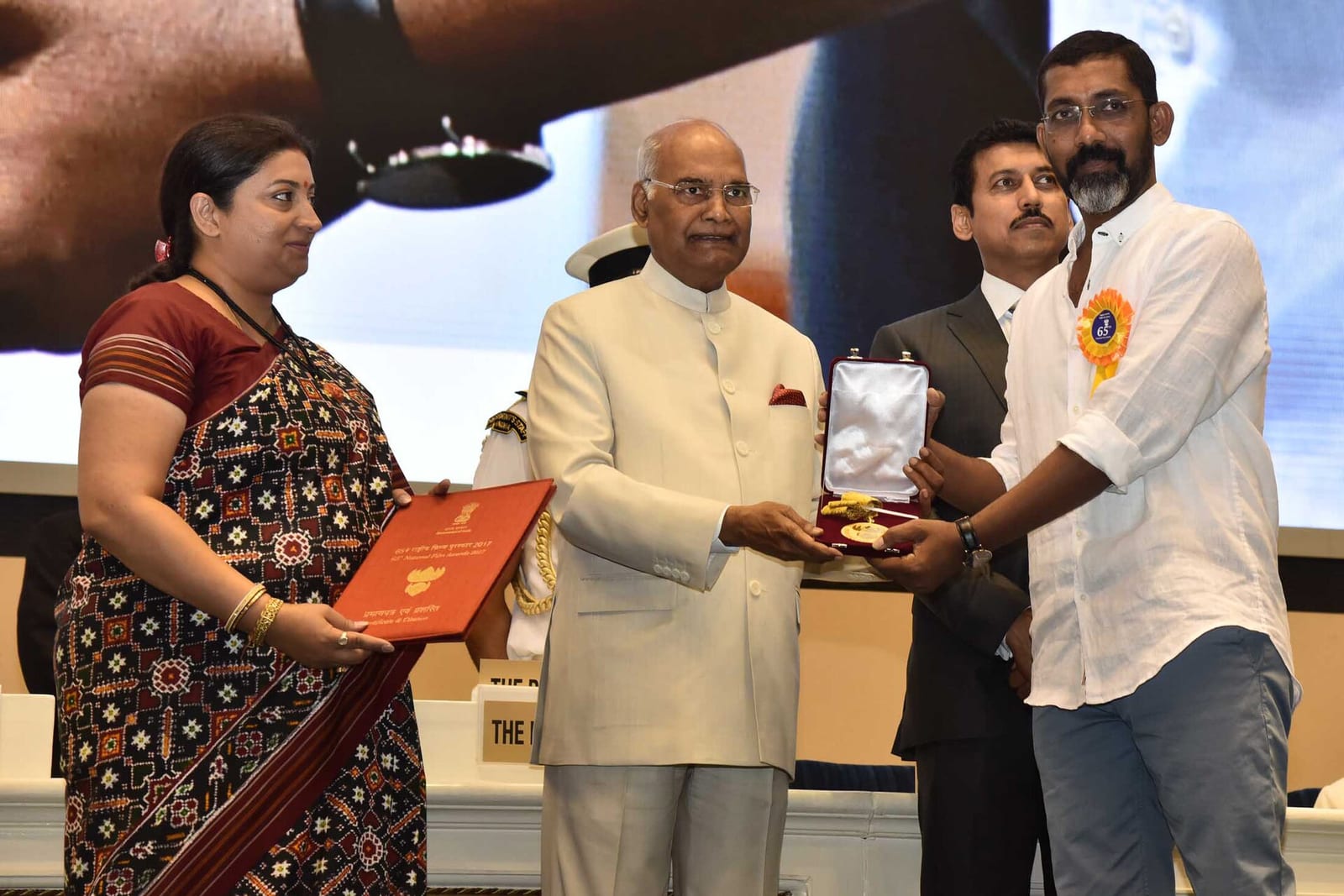 Nagraj Manjule, in a white shirt with a rosette, receives a medal in an open case from President Ram Nath Kovind on stage, with two ministers beside them