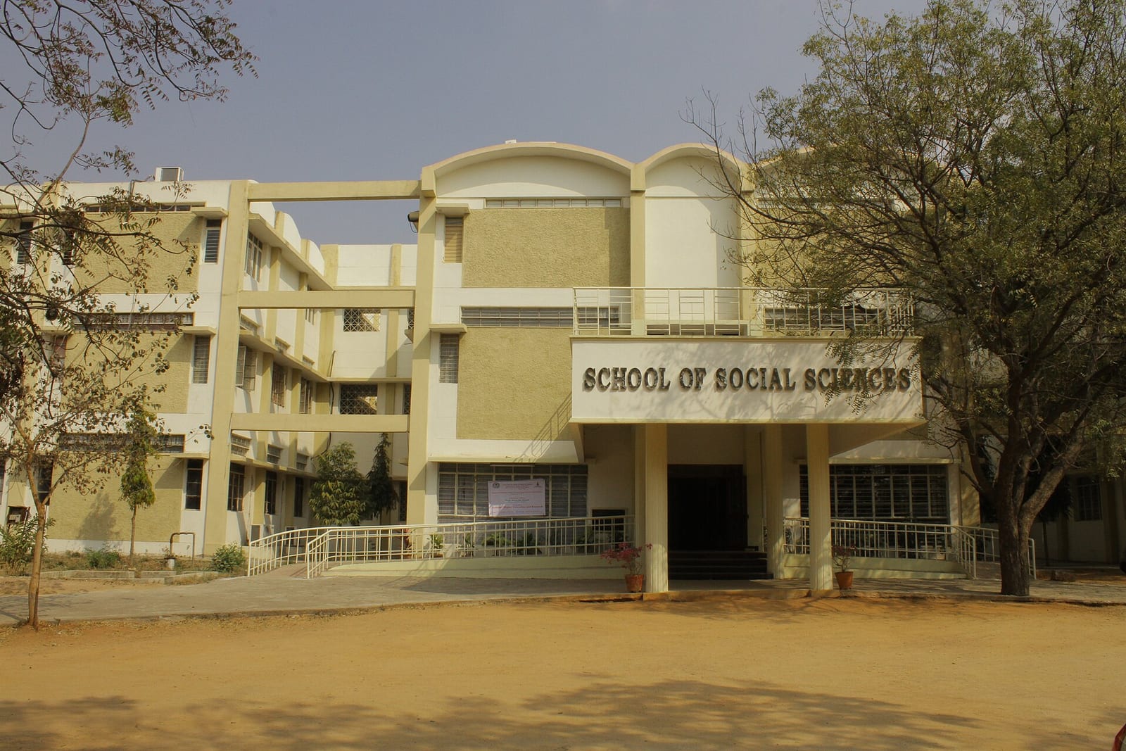 A cream-coloured multi-storey building with a 'School of Social Sciences' sign over its entrance, behind trees and a dirt forecourt