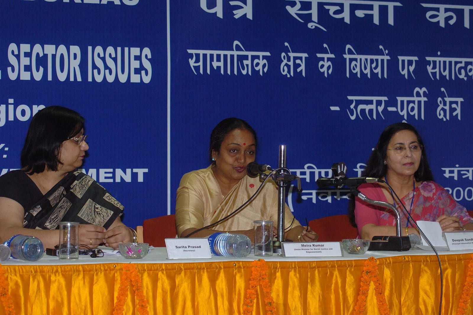 Meira Kumar, in a cream sari, speaks into a microphone at a conference table between two women officials
